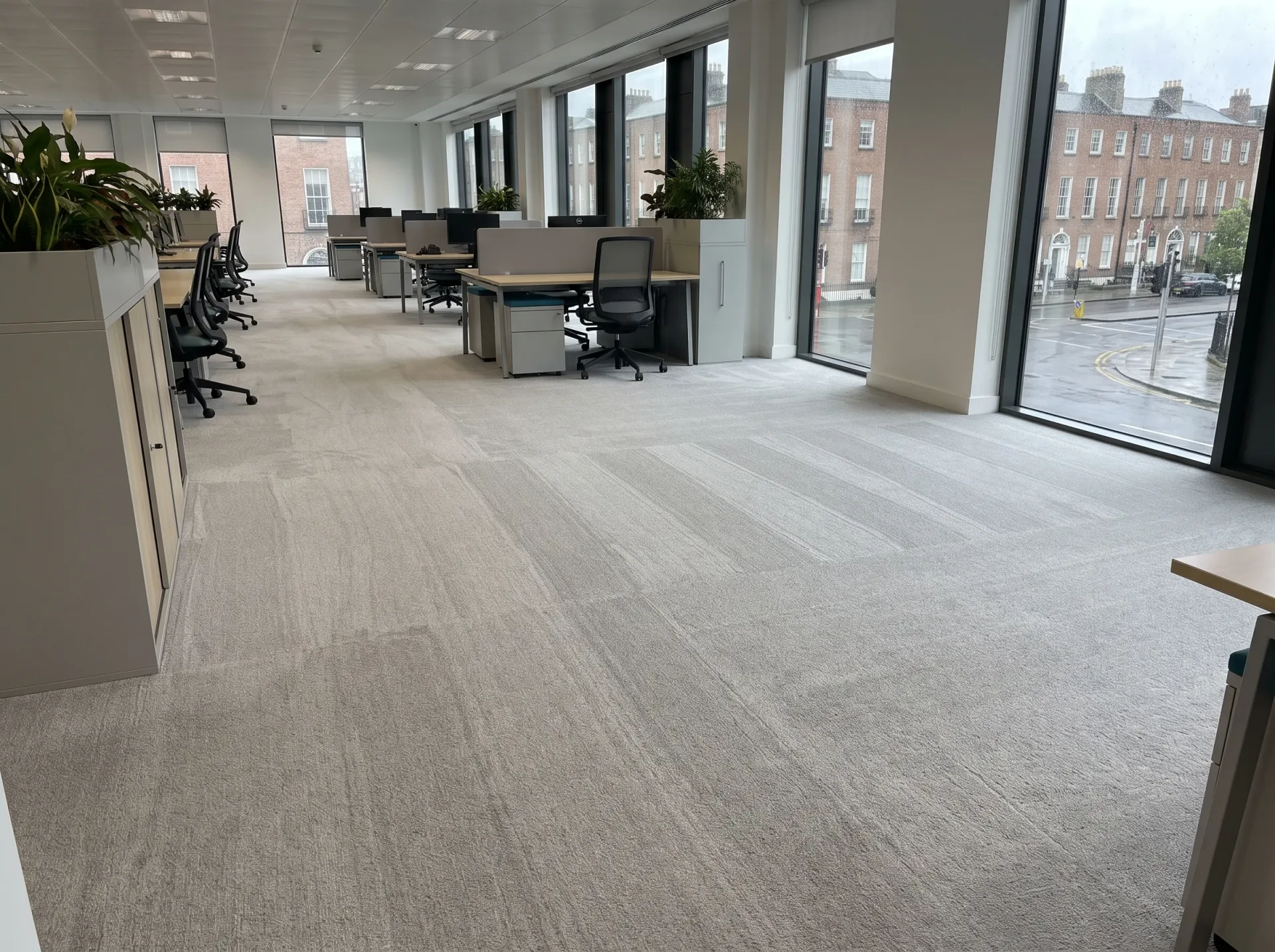 Spacious modern office with light-colored commercial carpet flooring, professional workstations, and large floor-to-ceiling windows